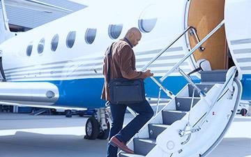Male business traveler with a laptop bag on his shoulder is looking at his smartpone while walking up the stps of a private jet