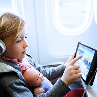 小女孩戴着耳机，腿上放着一个娃娃，在机上WiFi上玩平板电脑游戏